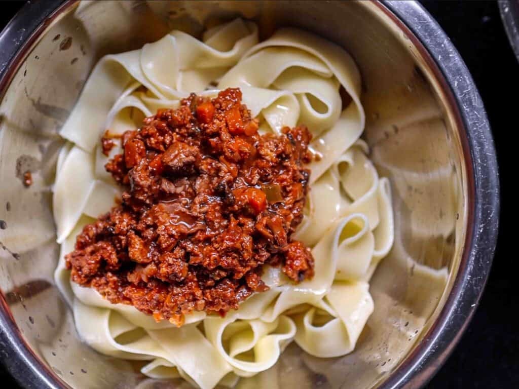 A bowl of wide, flat pasta noodles topped with a generous portion of hearty vegan bolognese sauce, featuring visible pieces of plant-based ground and diced vegetables.